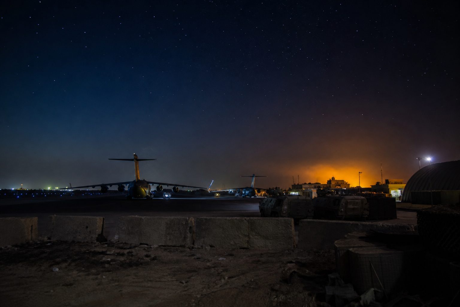 Military airbase perimeter at night with armored vehicles and runway lights in the distance.