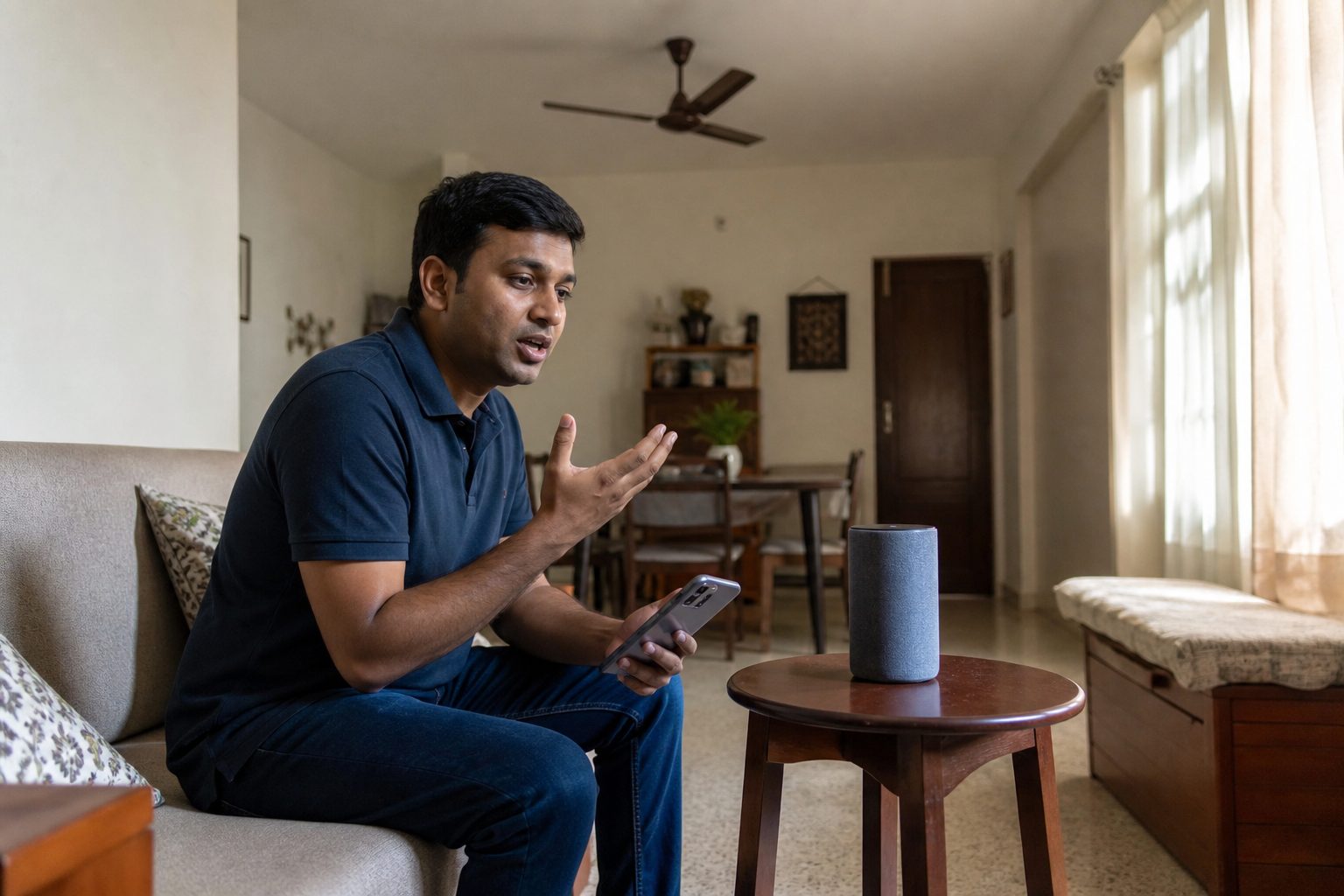 Person on a sofa speaking to a smart speaker with a phone in their lap in a modest Indian living room.