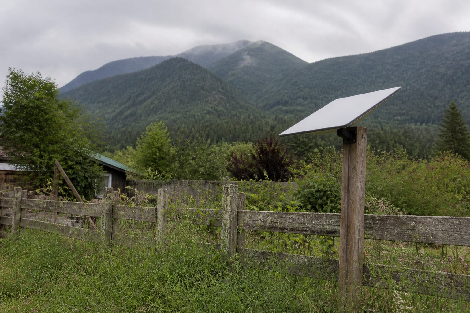 A flat rectangular satellite terminal mounted on a wooden post in a rural mountain backyard.