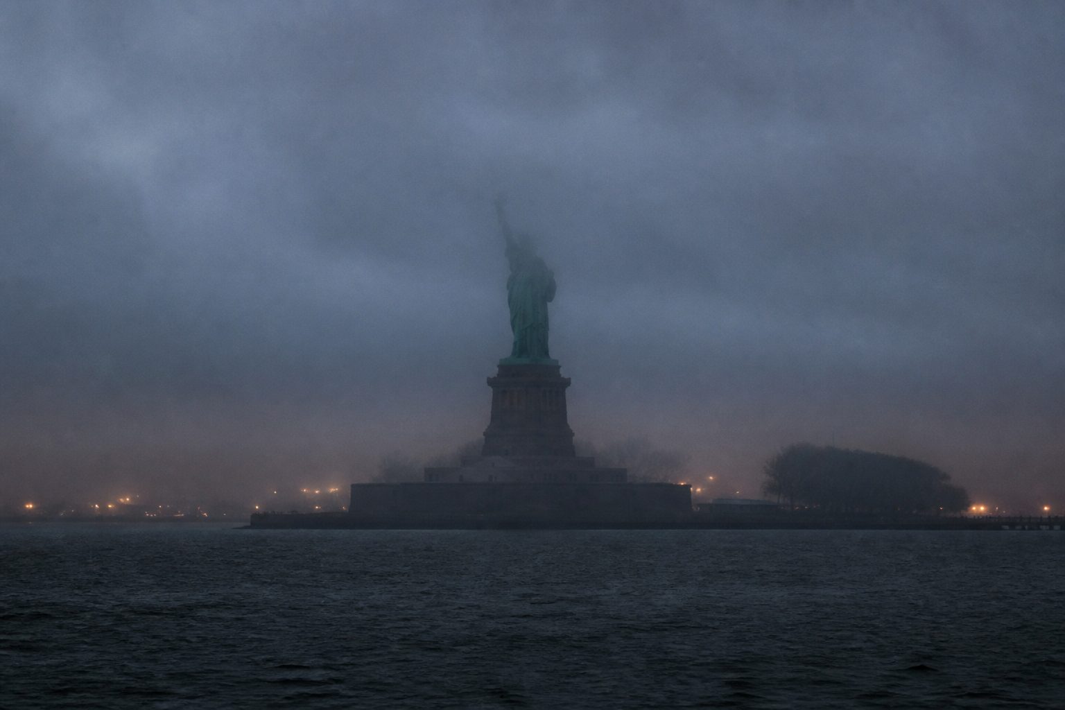 The Statue of Liberty shrouded in thick harbor fog at dusk, its torch obscured by mist.